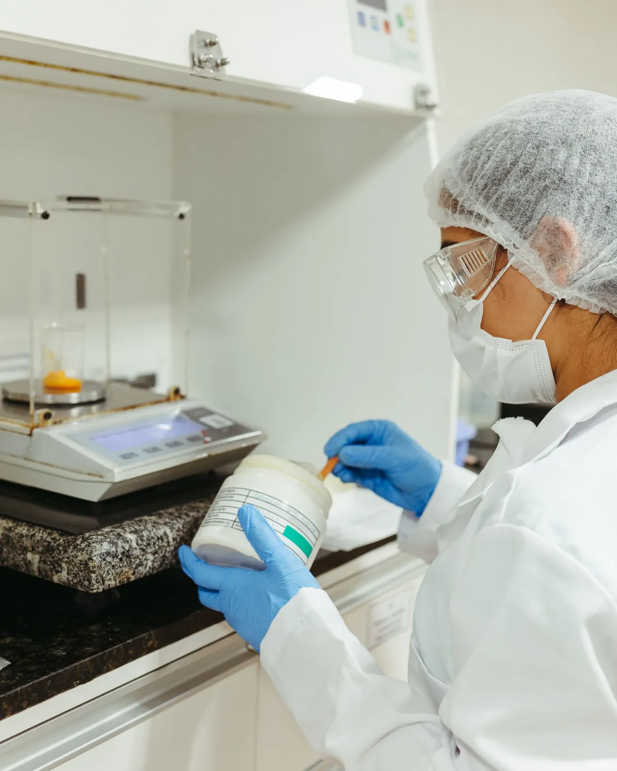A laboratory technician in protective gear taking a sample beside an analytical balance