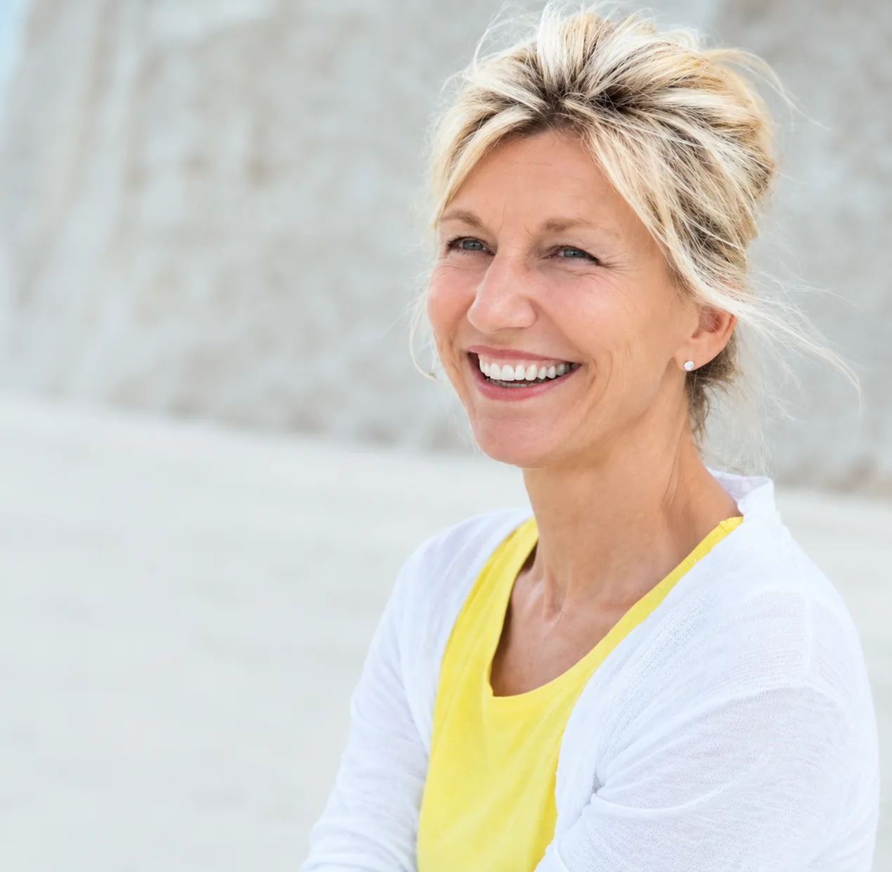 A smiling woman in a white cardigan and yellow top outdoors in soft daylight